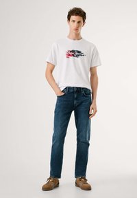 Camiseta de algodón blanca con diseño gráfico en texto, combinada con jeans de mezclilla azul oscuro y zapatillas marrones, con cordones y acentos sutiles.