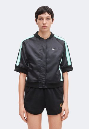 Άτομο που φοράει μαύρο μπουφάν Nike με κοντό μανίκι και ριγέ σε μέντα, καθώς και μαύρα σορτς, στέκεται μπροστά σε ένα απλό φωτεινό φόντο.