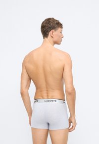 Grå bomullsboxere med svart kant som har påskriften "LACOSTE." Modellen vises bakfra, noe som viser en tettsittende design.