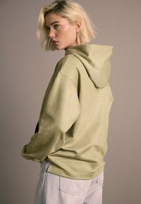 Olivovo zelená mikina s kapucňou z mäkkej tkaniny, vybavená šnúrkou na kapucni, dlhými rukávmi a voľným strihom. Dopĺňajú ju svetlosivé nohavice.