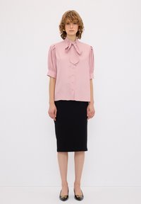 Blusa rosa con fiocco, maniche a palloncino corte, chiusura con bottoni. Abbinata a una gonna a tubino nera e scarpe nere con dettagli dorati.