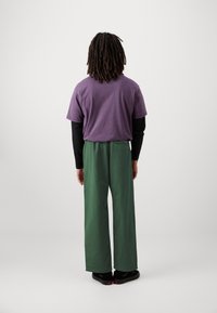 Paarse t-shirt over een zwarte lange mouwen shirt; losse groene broek; zwarte schoenen; zichtbaar gekruld haar aan de achterkant.