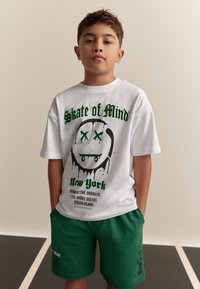 Wit katoenen T-shirt met een groene grafische afbeelding van een smiley en de tekst "Skate of Mind New York." Gecombineerd met groene korte broek.