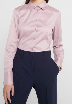 Femme portant une chemise rose pâle à manches longues rentrée dans un pantalon taille haute bleu marine foncé, une main dans la poche.