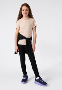 Jauna mergaitė su ilgais garbanotais plaukais, vilkinti šviesiai rudo Champion marškinėlius, juodas leggings, juodą striukę apjuosusi juosmenį ir juodai-purpurinius sportbačius.