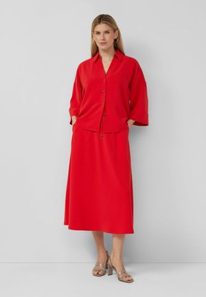 Frau steht mit den Händen in den Taschen, trägt eine rote Bluse mit Knöpfen und weitem Ärmel sowie einen passenden Midirock, kombiniert mit beigen Sandaletten mit Absatz.