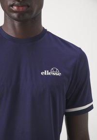 Námořnická modrá sportovní tričko s krátkým rukávem, s bílými akcenty na manžetách a bílým logem "ellesse" na hrudi.