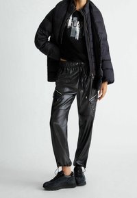 Sort pufferjakke, sort grafisk t-shirt og skinnende sorte joggers i faux læder med lynlåslommer; sorte sneakers med teksturerede detaljer.
