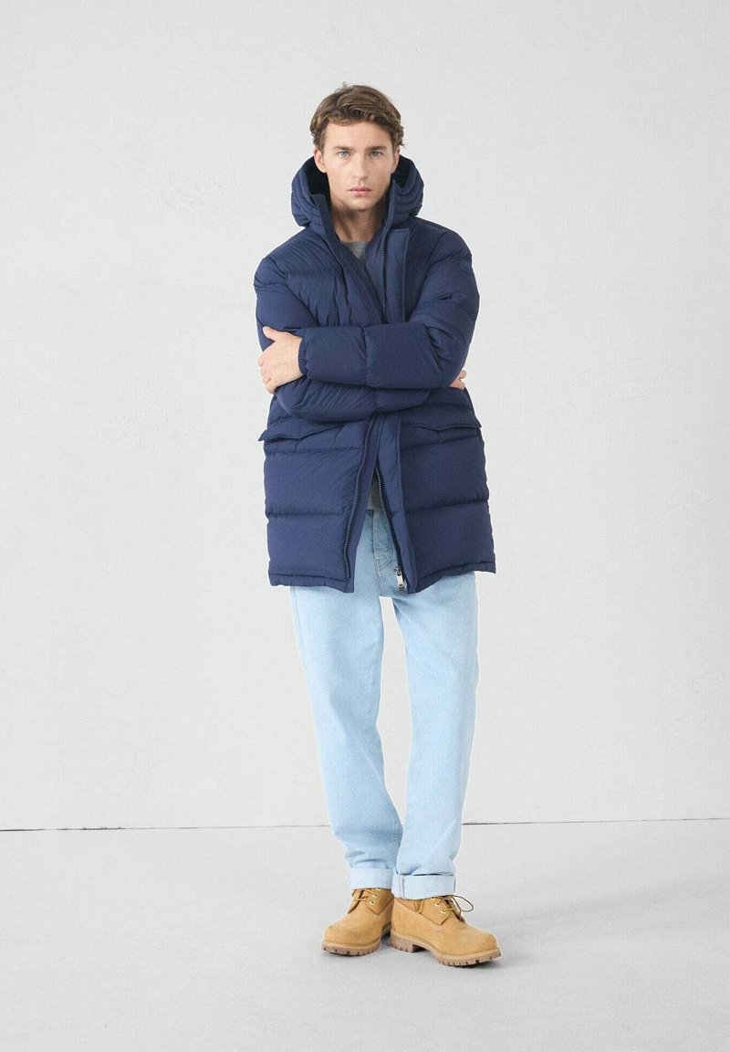 Jeune homme portant une doudoune bleu marine, un jean bleu clair et des bottes beiges à lacets, debout avec les bras croisés devant un fond uni.