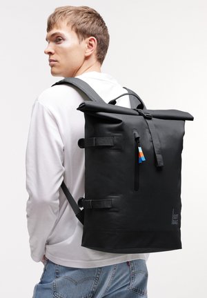 Joven con camisa blanca de manga larga y vaqueros azules que lleva una gran mochila negra con cierre enrollable, correas laterales y bolsillo delantero con cremallera.