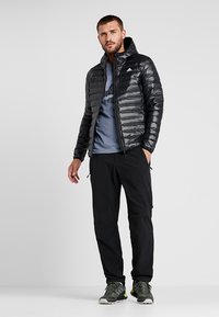 Schwarze isolierte Jacke mit einem gesteppten Design, kombiniert mit einem grauen T-Shirt und schwarzen Hosen. Modell trägt grau-schwarze Sportschuhe.