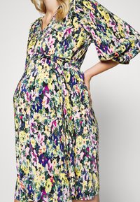 Vestido de maternidade com padrão floral, apresentando mangas curtas e bufantes, decote em V e cintura com cinto, nas vibrantes cores amarelo, rosa e azul.