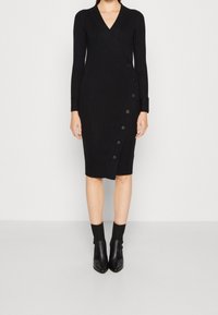 Donna che indossa un abito midi a coste nero con scollatura a V, maniche lunghe, dettagli con bottoni laterali, calze nere alla caviglia e scarpe nere con punta appuntita.
