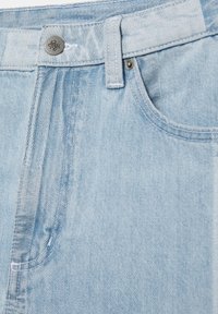 Jean en denim bleu clair en gros plan montrant la taille, le bouton, la passants de ceinture, la poche avant et les détails des rivets métalliques.