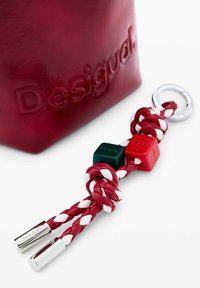 Punane nahktasku, millel on reljeefne "Desigual" logo, esitleb punast, valget ja rohelist punutud nööri ning metallist aksessuaare.