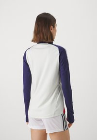 Camisa deportiva de manga larga en blanco y azul marino con acentos rojos en contraste. Fabricada con una tela suave y ligera. Presenta un diseño ajustado.