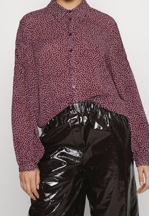 Persona con una camiseta de manga larga color burdeos con puntos blancos metida en unos pantalones de cuero patentado negro de cintura alta y brillantes.