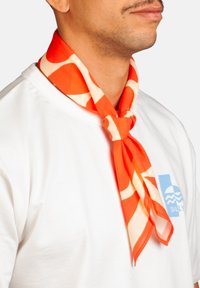 Homme portant un t-shirt blanc avec un logo bleu et une écharpe orange à motifs nouée autour de son cou, sur un fond uni.