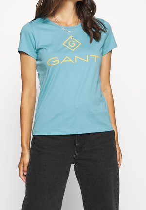 Sinihtävänvihreä puuvillainen t-paita, jossa on lyhyet hihat ja pyöreä pääntie. Edessä suuri keltainen "GANT"-logo timantin muodossa.