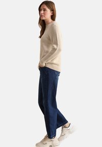 Beige gebreide trui met een textuurpatroon, gecombineerd met donkerblauwe rechte jeans en lichtgekleurde sneakers.