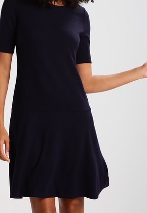 Femme portant une robe bleu marine foncé à manches courtes, longueur genou, sur un fond clair uni, bras tendu sur le côté.