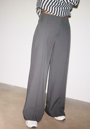 Osoba stojaca na betónovej podlahe pri bielej stene, oblečená v sivých širokých nohaviciach s prednými záhybmi a bielych teniskách.