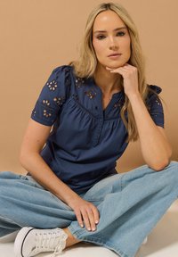 Top azul marino de manga corta con diseños de flores recortadas, tapeta de botones y detalles fruncidos, combinado con unos vaqueros de pierna ancha de color azul claro.