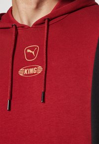 Felpa rossa con cappuccio e pannelli laterali neri, con logo Puma dorato e testo "KING". Realizzata in tessuto morbido con cordini regolabili.