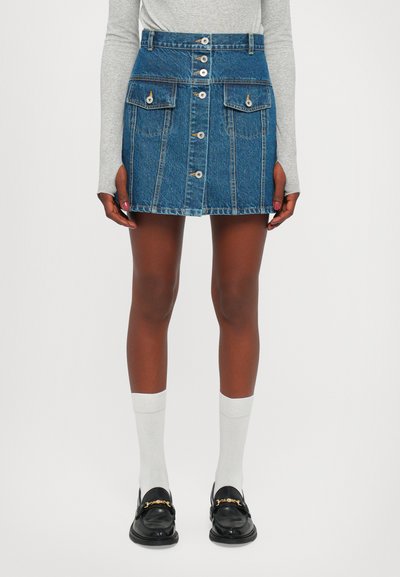 Denimnederdel med tætsiddende talje, fem-knaps lukning, forlommer og blå vask. Bæres med grå lange ærmer, hvide sokker og sorte slip-on sko.