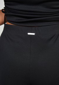 Černý crop top z žebrované tkaniny se shrnovacími detaily a malou stříbrnou visačkou na zadním horním švu, který ukazuje texturovaný vzhled.