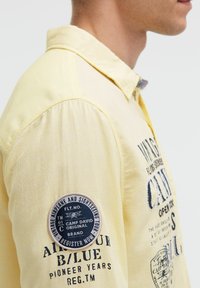 Gelbes Hemd mit Knopfleiste und strukturiertem Finish, mit einem Patch am Ärmel und bedruckten Grafiken auf der Vorderseite.