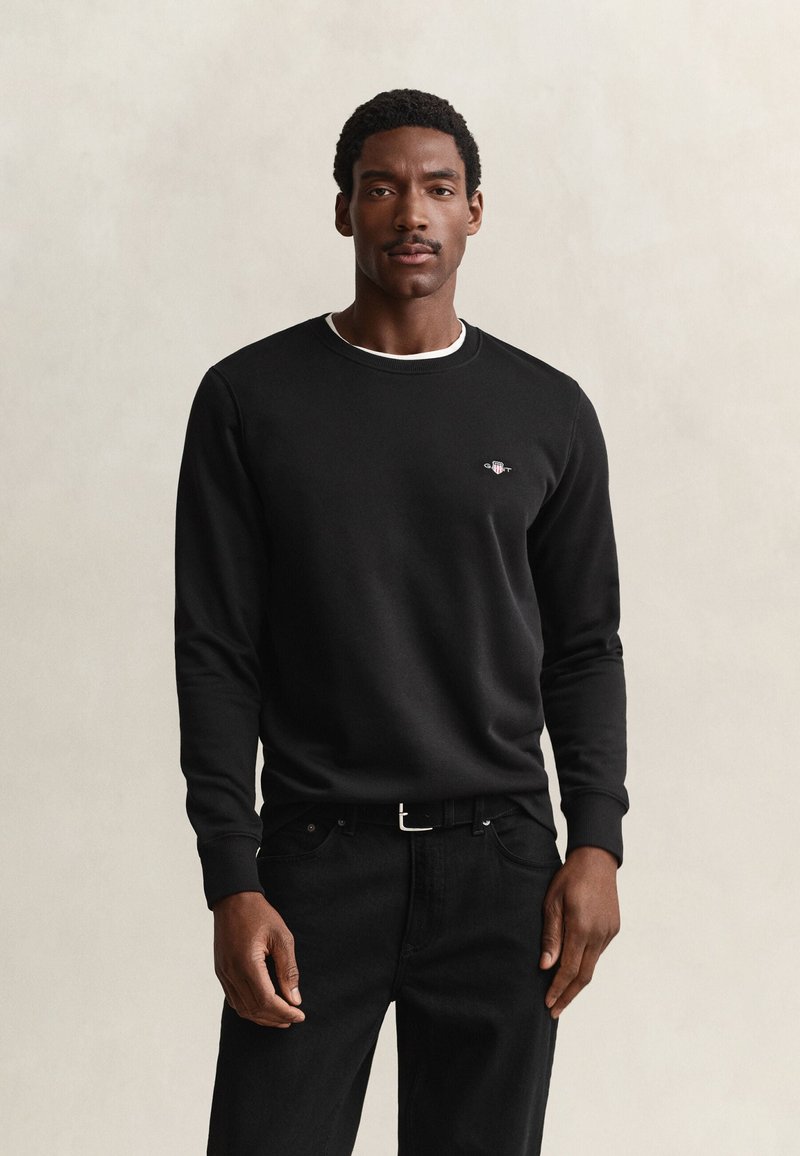Homme portant une chemise noire à manches longues et un pantalon noir, debout devant un fond clair uni, regardant devant avec une expression neutre.