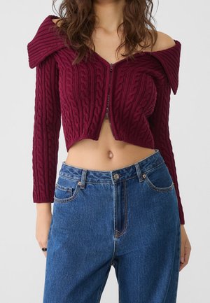 Kvinde iført en bordeaux off-shoulder kabelstrikket cropped sweater og højtaljede blå jeans, som viser maven og navlen.
