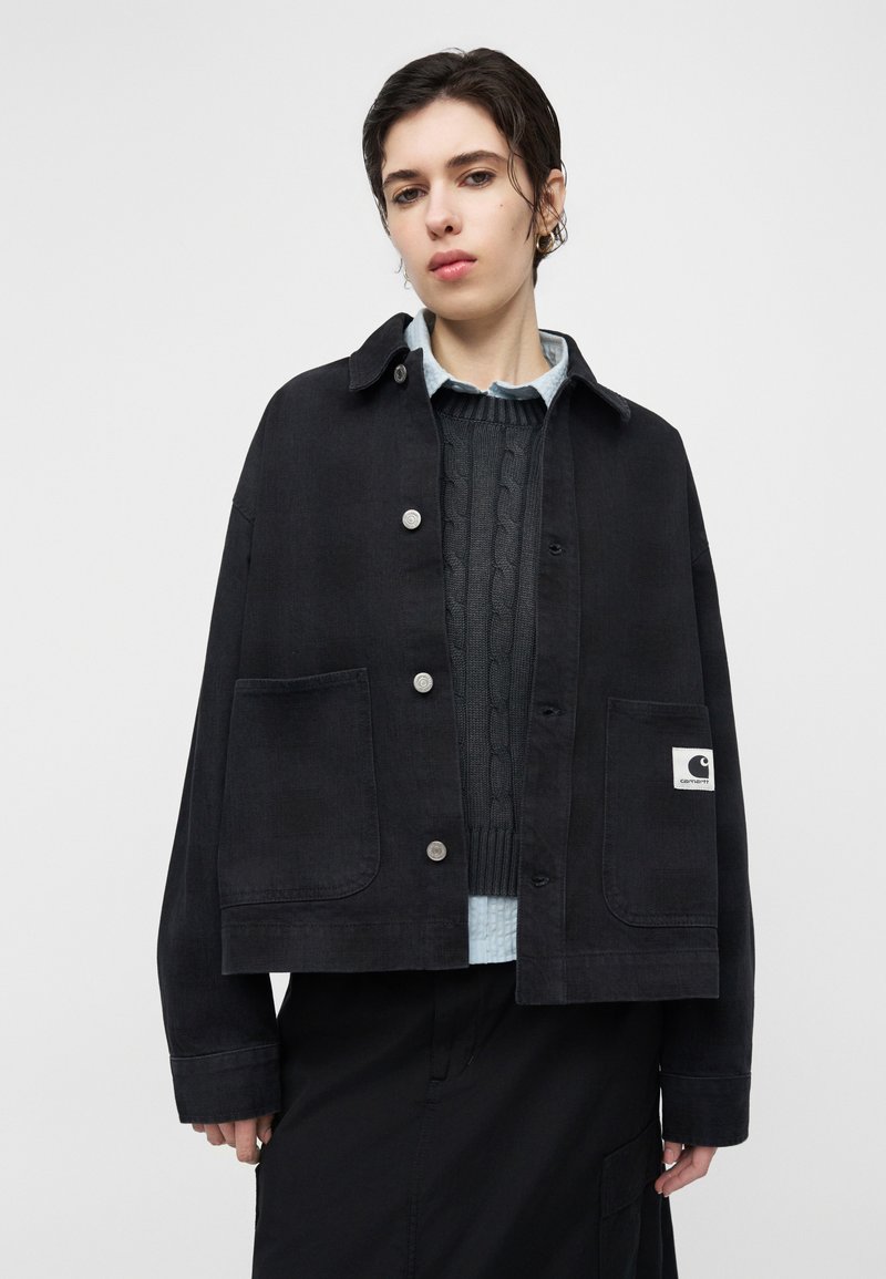 Person wearing a black button-up jacket with large pockets over a dark cable-knit sweater and light blue collared shirt.