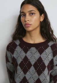 Joven con el pelo largo y oscuro, vistiendo un suéter con patrón de rombos en tonos burdeos y gris, de pie frente a un fondo claro y liso.