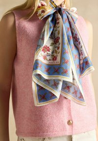 Růžový svetřík bez rukávů s texturovaným povrchem, doplněný modro-bílým hedvábným šátkem s květinovým vzorem uvázaným kolem krku. Detail zlatých knoflíků.