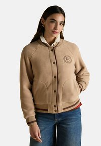 Bež flanellist jakk, millel on pruunid aksessuaarid, ümar kaelus ja soonikkoes varrukad. Omab logoga täket ja kahte esist pocketit.