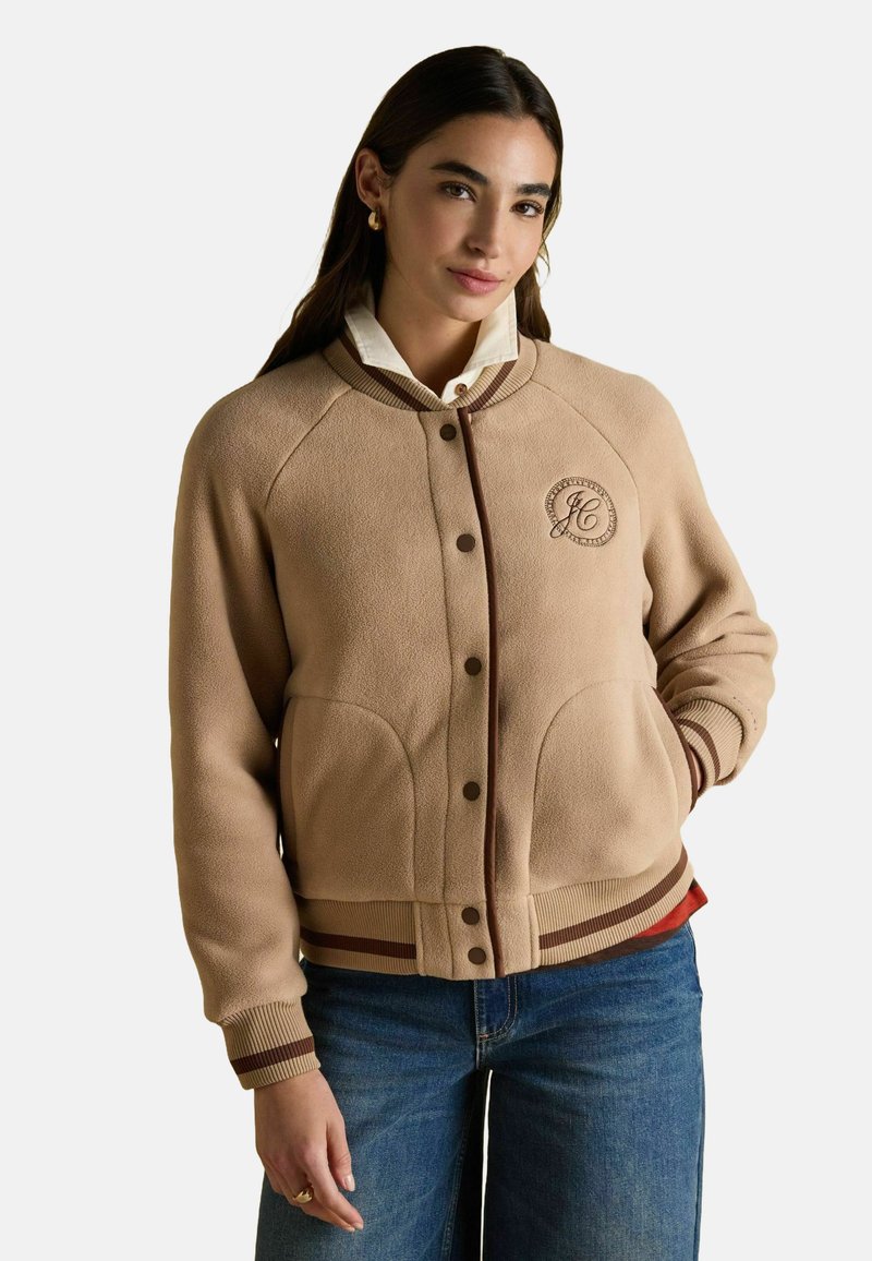 Bež flanellist jakk, millel on pruunid aksessuaarid, ümar kaelus ja soonikkoes varrukad. Omab logoga täket ja kahte esist pocketit.