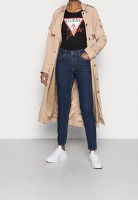 Person in beigem Trenchcoat, schwarzem Guess-Logo-T-Shirt, blauen Jeans und weißen Sneakers, steht mit einer Hand in der Tasche.