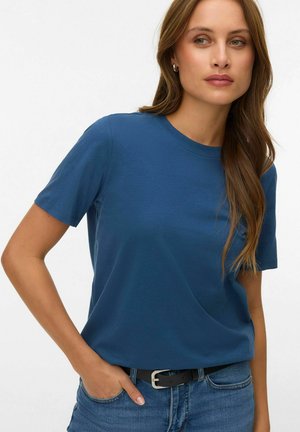 Woman with long brown hair wearing hoop earrings, a blue short-sleeve shirt, and blue jeans with a black belt, standing with one hand in pocket.