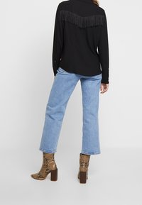 Chemise noire boutonnée avec des détails à franges au dos, associée à un jean droit en denim bleu clair et des bottines marron en peau de serpent.