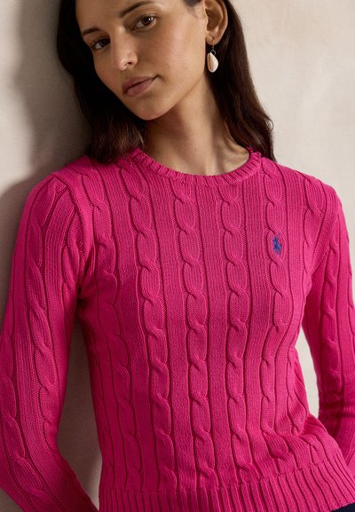 Vrouw draagt een felroze kabelgebreide trui met een klein blauw logo, staand tegen een eenvoudige lichte achtergrond.