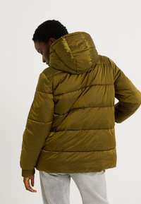 Doudoune vert olive avec capuche, présentant un motif cousu et des poignets côtelés. Matière lisse et brillante avec une coupe décontractée.