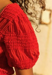 Rød bluse med sammenkrøllede, teksturerede puffærmer og smock-detaljer. Stof er letvægtigt med en glat overflade.
