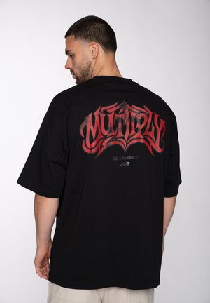 Man facing sideways wearing black oversized t-shirt with red stylized "MULTIPLY" text on the back, beige pants, and a short haircut.