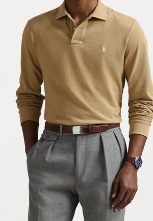 Homme portant un polo à manches longues beige, un pantalon plissé gris, une ceinture en cuir marron et une montre-bracelet avec un cadran bleu et un bracelet marron.
