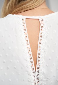 Witte blouse met een gestructureerde stof met kleine gestippelde patronen, een gegolfde kanten rand en een keyhole-achterzijde.