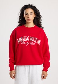 Rode sweatshirt met witte tekst "MORNING ROUTINE Varsity Club". Heeft een relaxte pasvorm, ronde hals en ribgebreide manchetten, gemaakt van zacht materiaal.