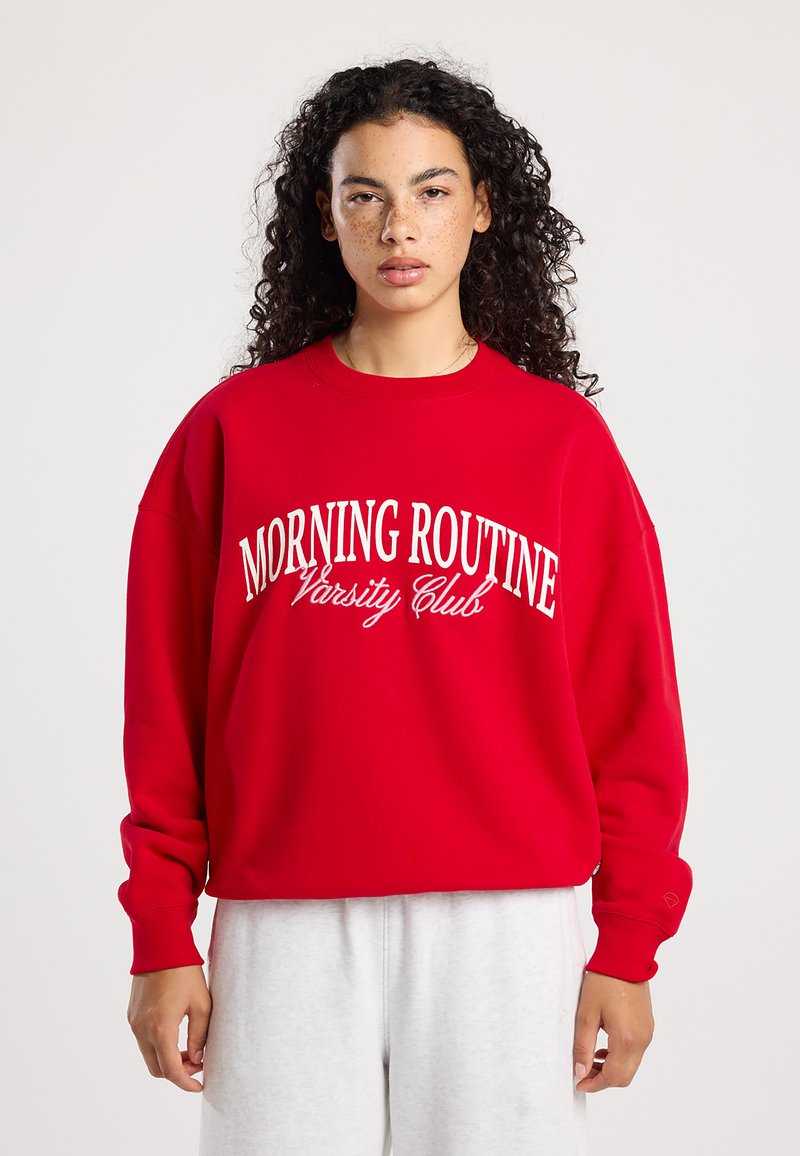 Rode sweatshirt met witte tekst "MORNING ROUTINE Varsity Club". Heeft een relaxte pasvorm, ronde hals en ribgebreide manchetten, gemaakt van zacht materiaal.