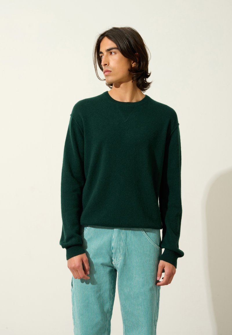 Grüner Strickpullover mit Rundhalsausschnitt und gerippten Bündchen, kombiniert mit hellblauen und weißen gestreiften Hosen, die einen lockeren Sitz zeigen.